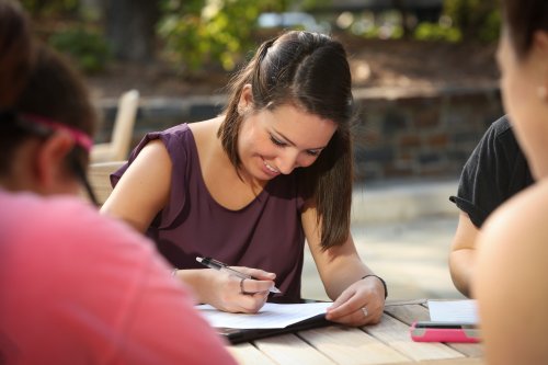 duke student sitting outside on a sunny day smiling and doing schoolwork.