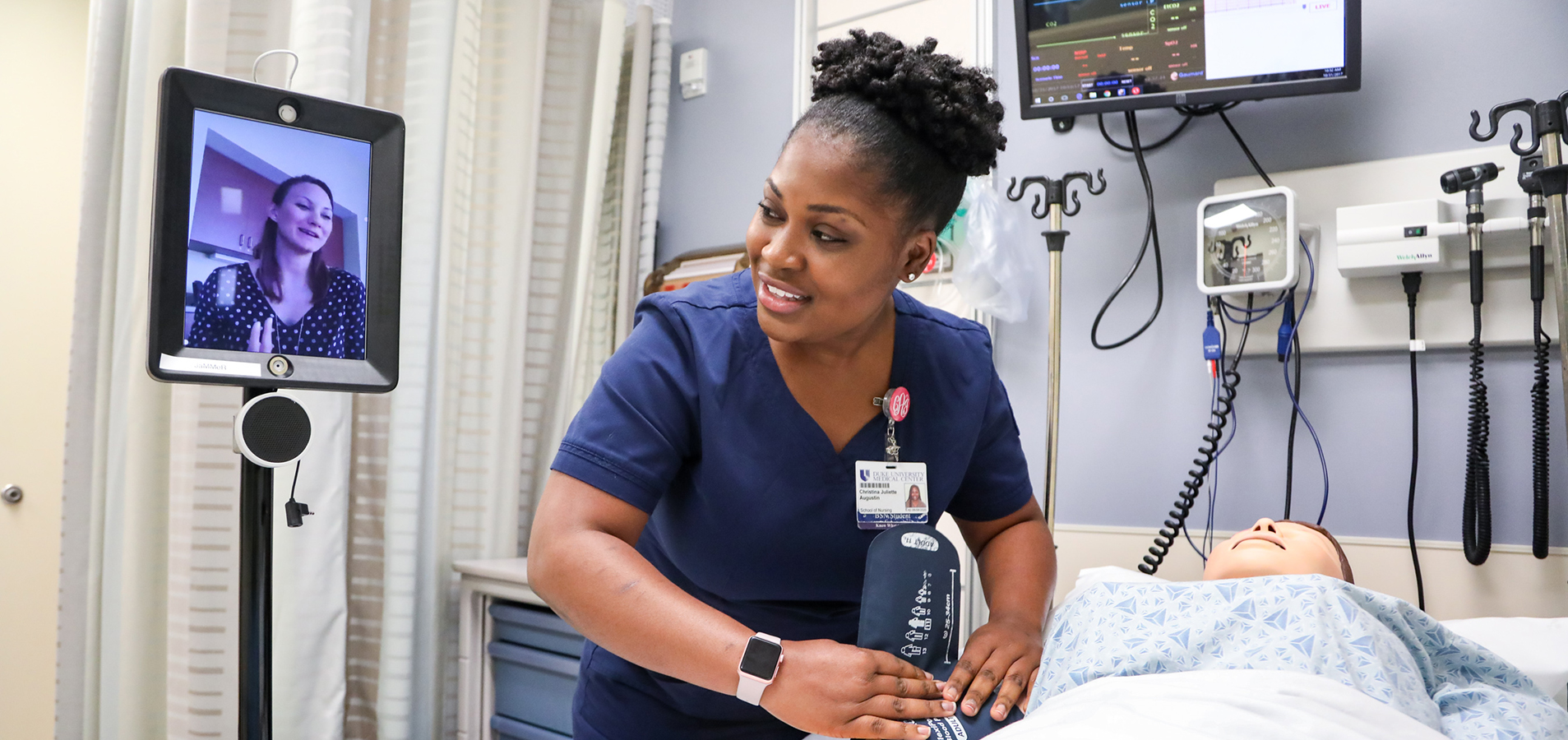 A female Duke University School of Nursing BSN student wearing blue scrubs leans over a hospital bed that contains a high-fidelity mannequin in a simulated critical care environment. An iPad sits next to her with faculty directing her in the simulated nursing experience.