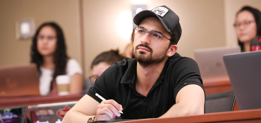 A male Duke University School of Nursing MSN student wearing a black polo shirt and a black hat sits in a sizeable lecture-style classroom and listens intently during intensives.