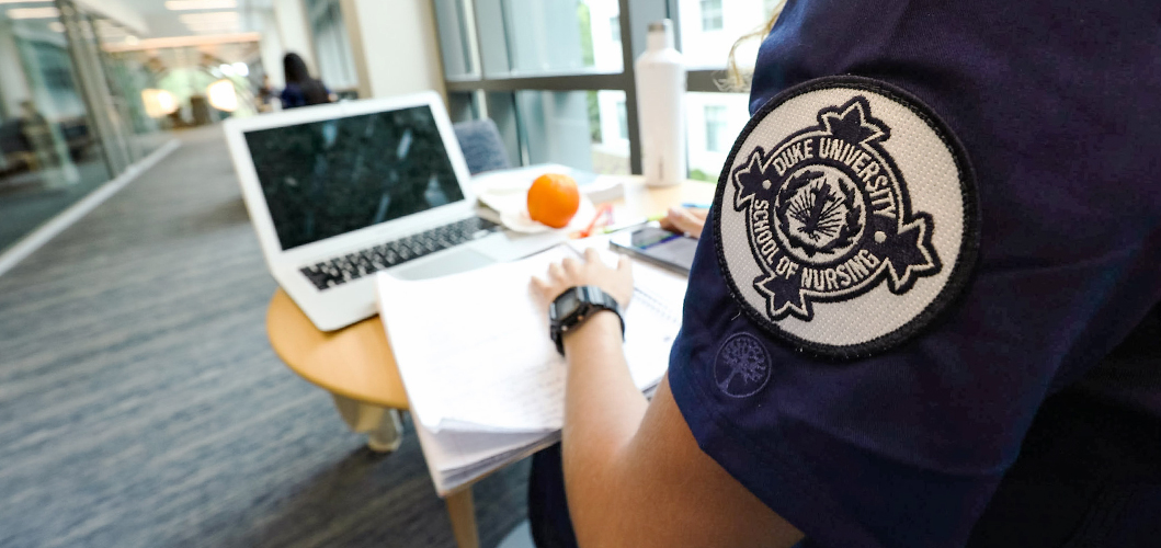 A female Duke University School of Nursing ABSN student studies and sits at a small round table with papers, a laptop, an orange, and a water bottle. Her shoulder patch is visible and contains a round Duke School of Nursing emblem on a patch.