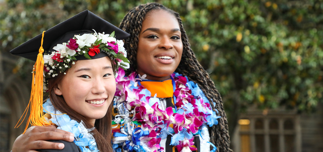 Two talented female Duke School of Nursing student classmates smile at a graduation ceremony. One student has her arm around the other and they both wear caps, gowns, and brilliant flowers around their necks.