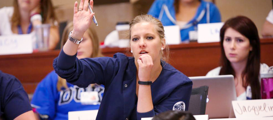 A Duke University School of Nursing Women's Health Care Nurse Practitioner student raises her hand and looks thoughtful among other students in a large classroom setting.