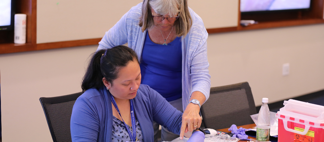 Kathy Trotter, a Duke University School of Nursing faculty member, leans over a Women's Health Nurse Practitioner student, smiles and guides the student on a technical skill.