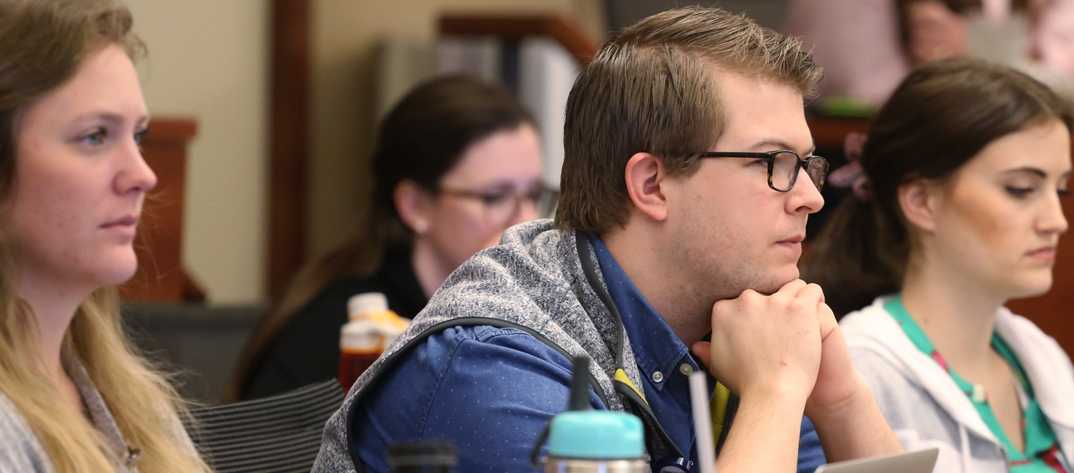 A male Duke University School of Nursing MSN Health Informatics student has his hands under his chin and looks thoughtful among other students in a large classroom setting.  