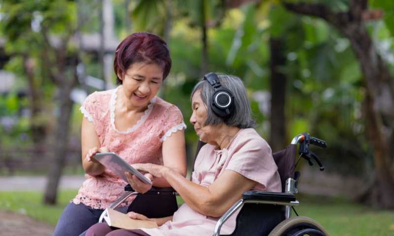 A younger Asian woman assists and older Asian woman in wheelchair with an iPad. The older woman is wearing headphones, and they are seated outdoors in a park-like setting.