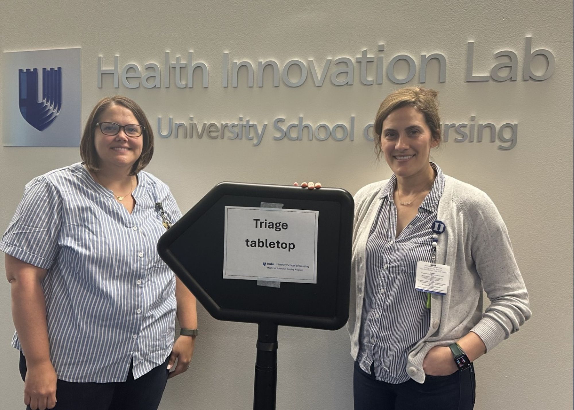 Brandi Snow and Laura Liggett with a sign pointing towards their Triage Tabletop simulation