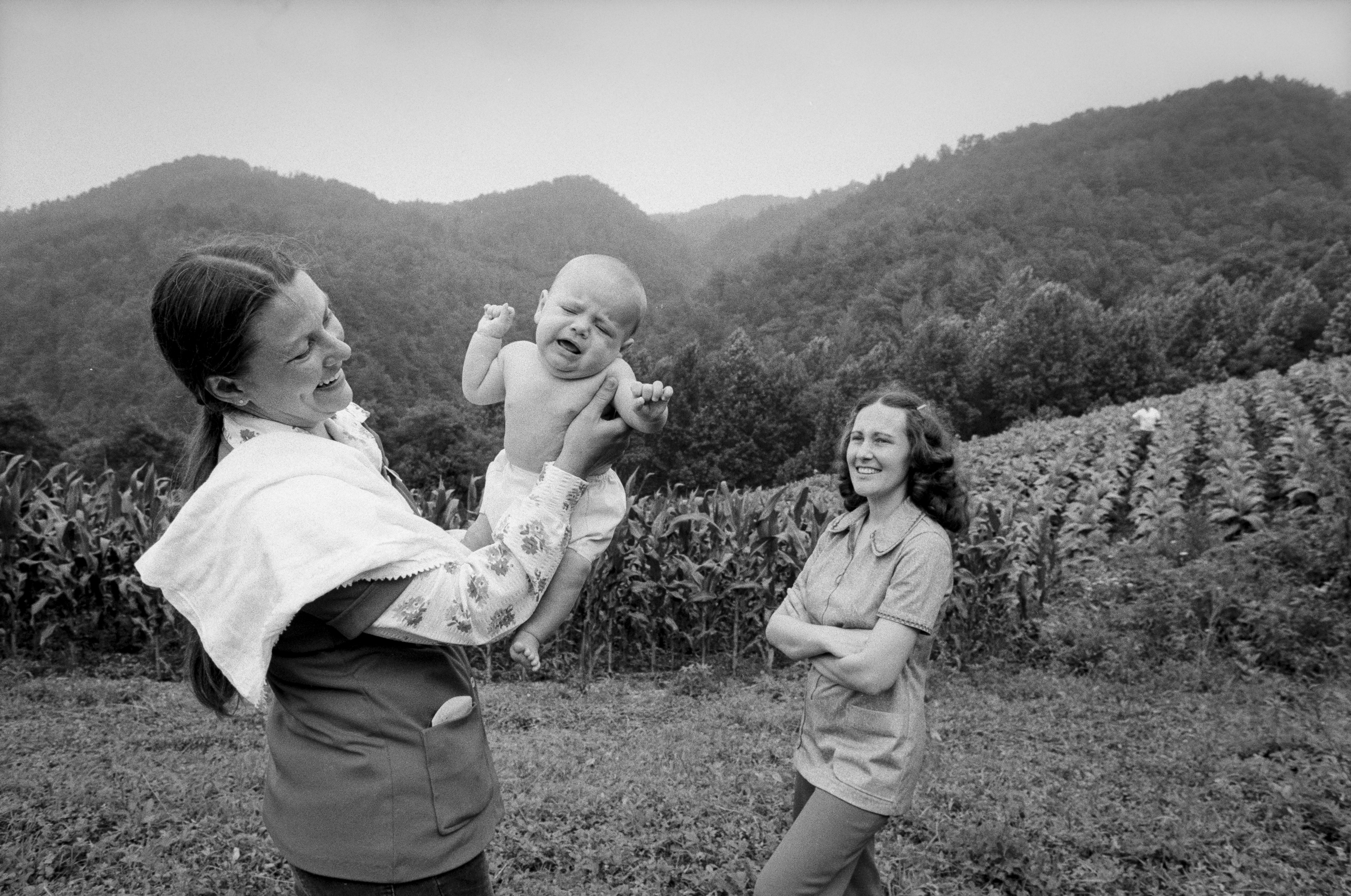 A black and white photo of a woman holding an infant while another woman looks on. In the background there are fields and mountains.