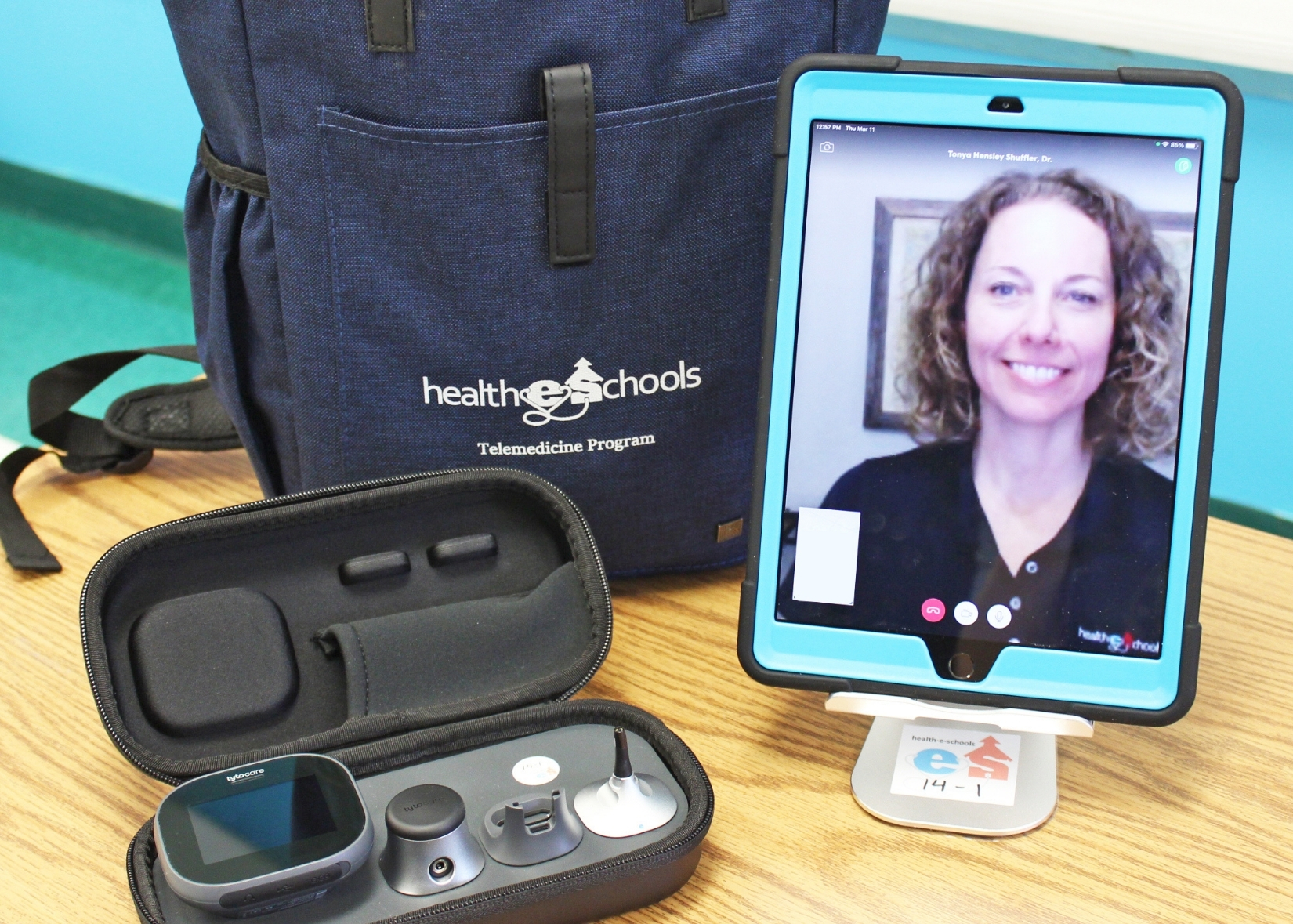 A nurse appears on a mobile screen as part of a telehealth visit. There is a backpack and audio equipment on the desk next to the tablet.