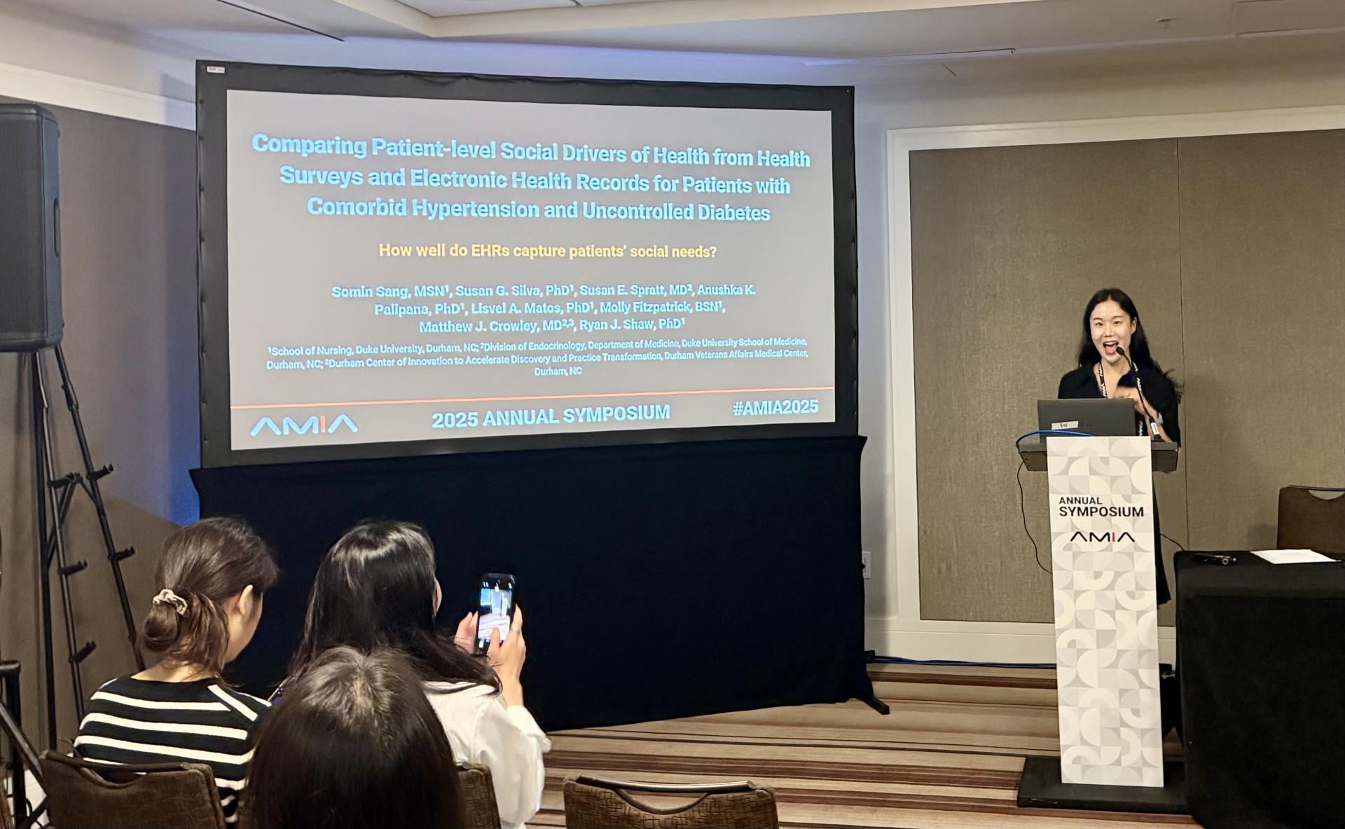A student stands at a podium giving a presentation, while a slideshow is displayed on a screen.