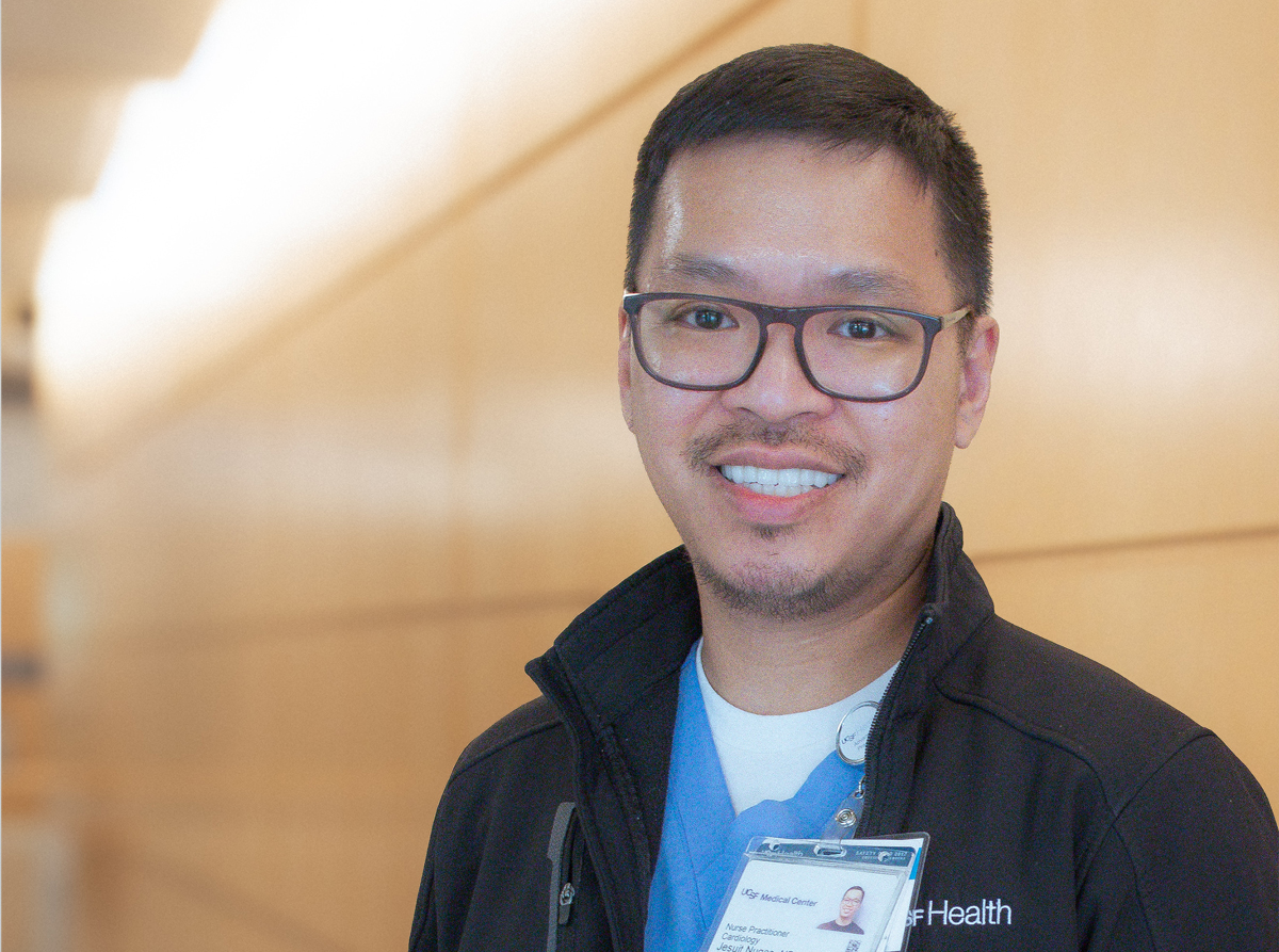 A nurse standing in a hallway wearing light blue scrubs and a dark jacket.
