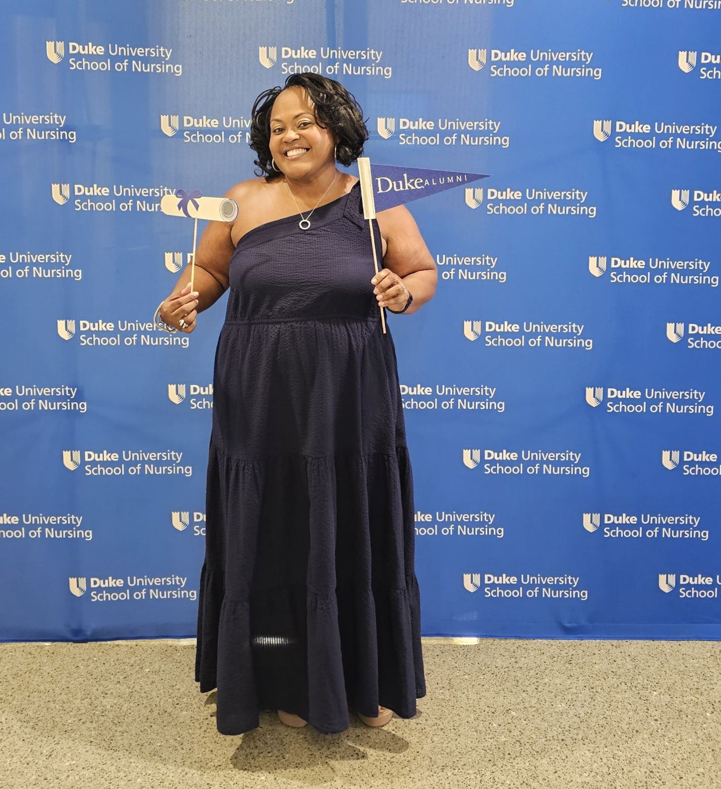 A person wearing a black gown poses in front of a Duke University School of Nursing backdrop.
