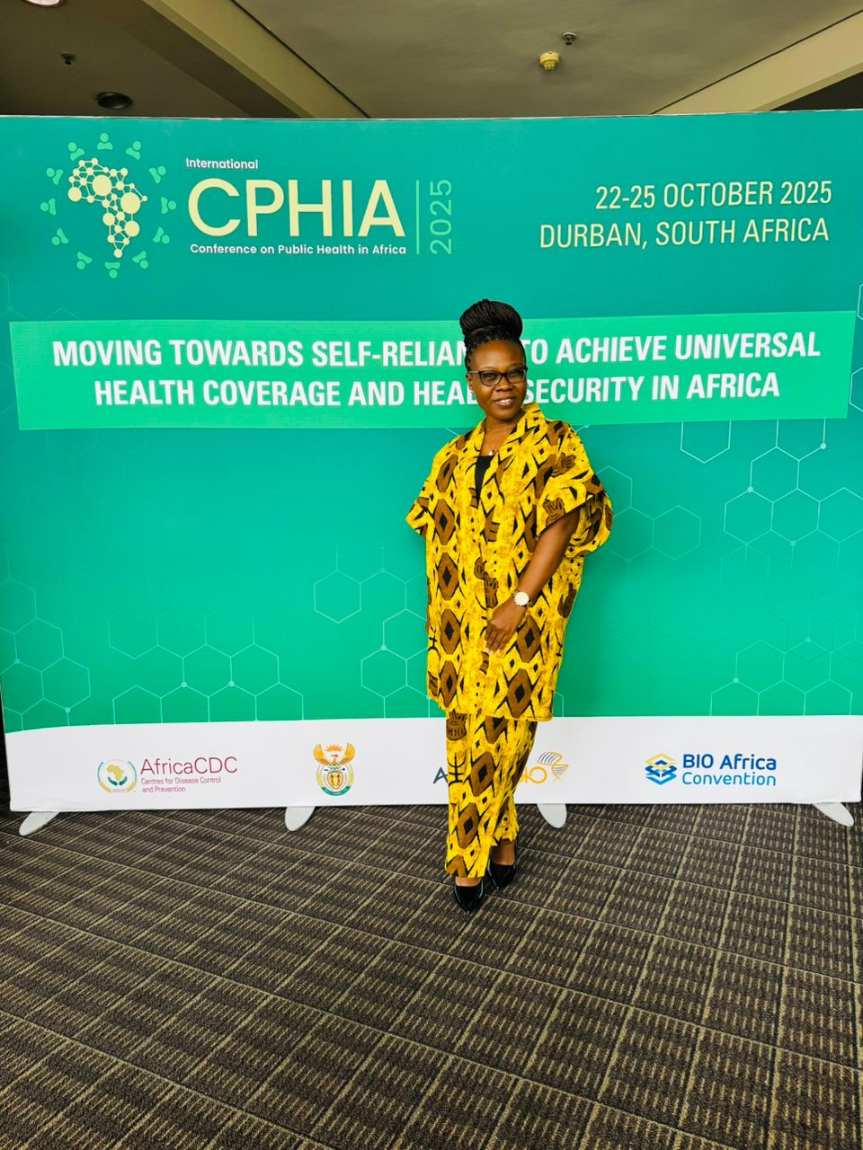 A woman in a bright yellow and patterned jumpsuit stands in front of a green background.