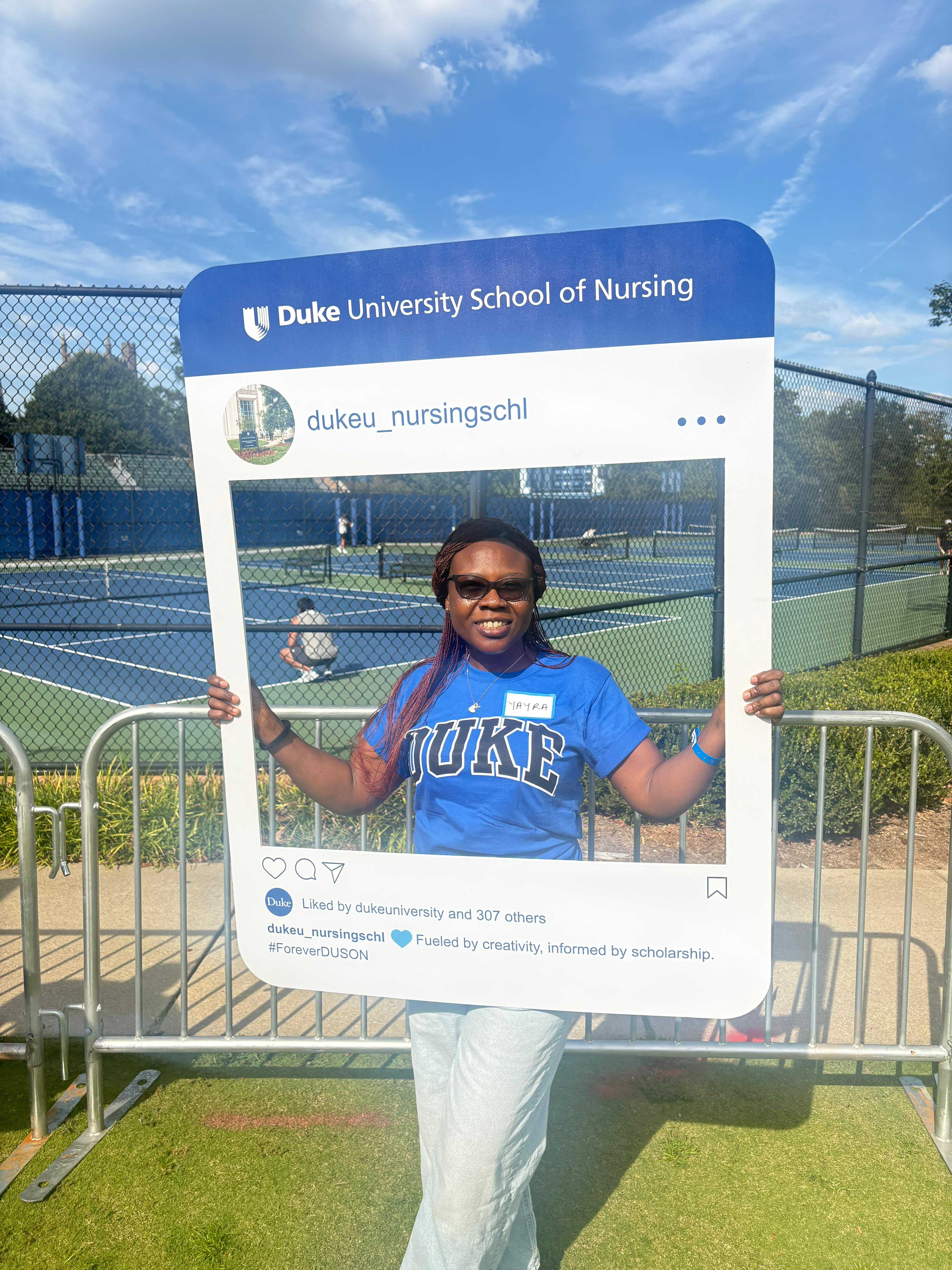 A woman in Duke blue stands behind a life-size Instagram frame, posing for a photo a Duke athletics event.