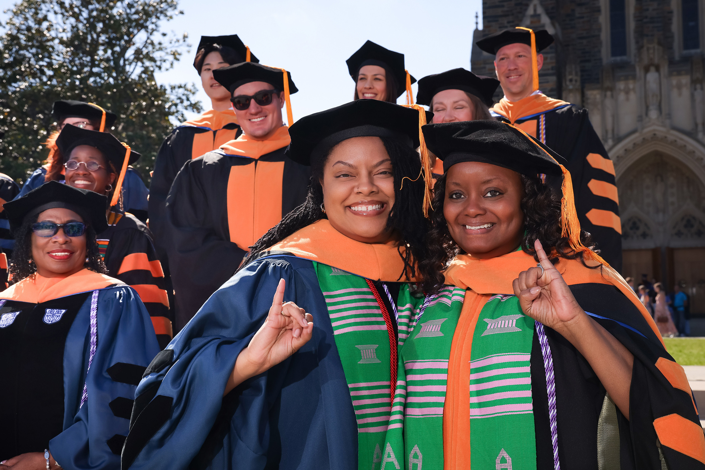 Two graduating students in cap and gown smile while other grads look on