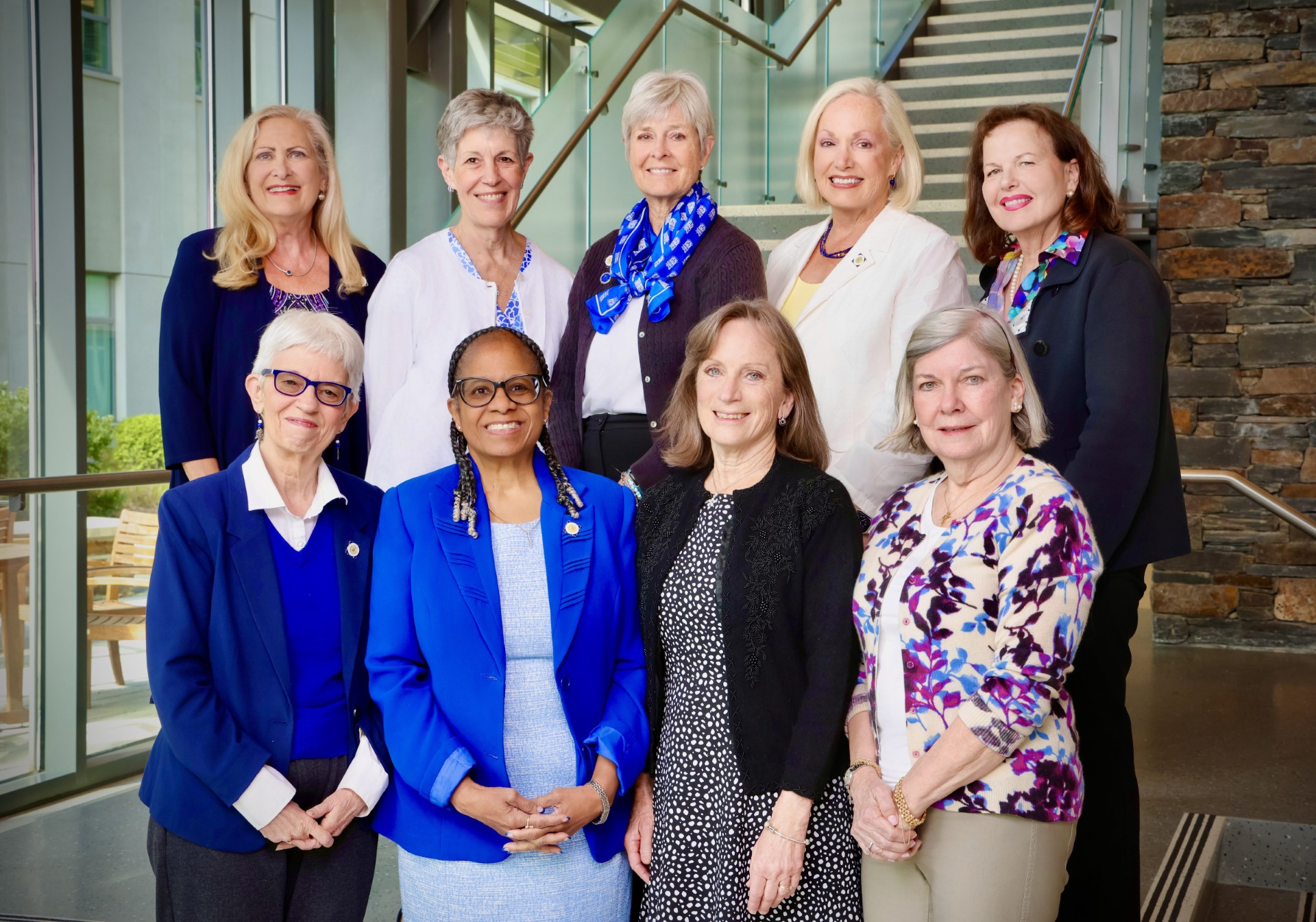 A group of nine women pose on a staircase in a well-lit atrium. Most are wearing some shade of blue.