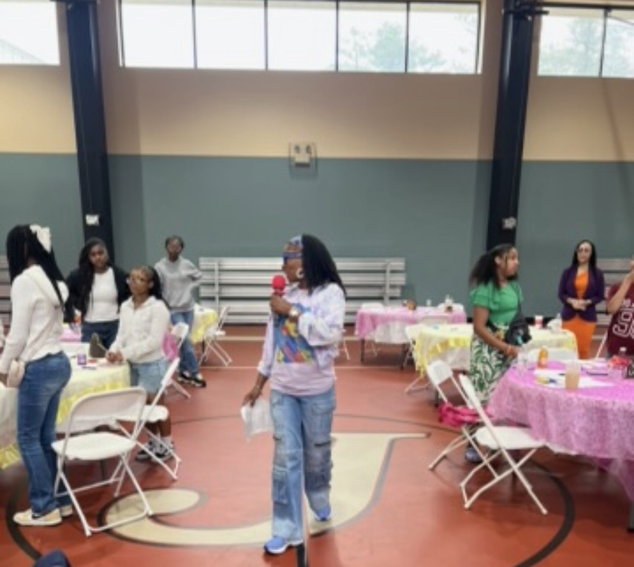 Lydia Nichols with a microphone in a school gymnasium with girls standing around tables