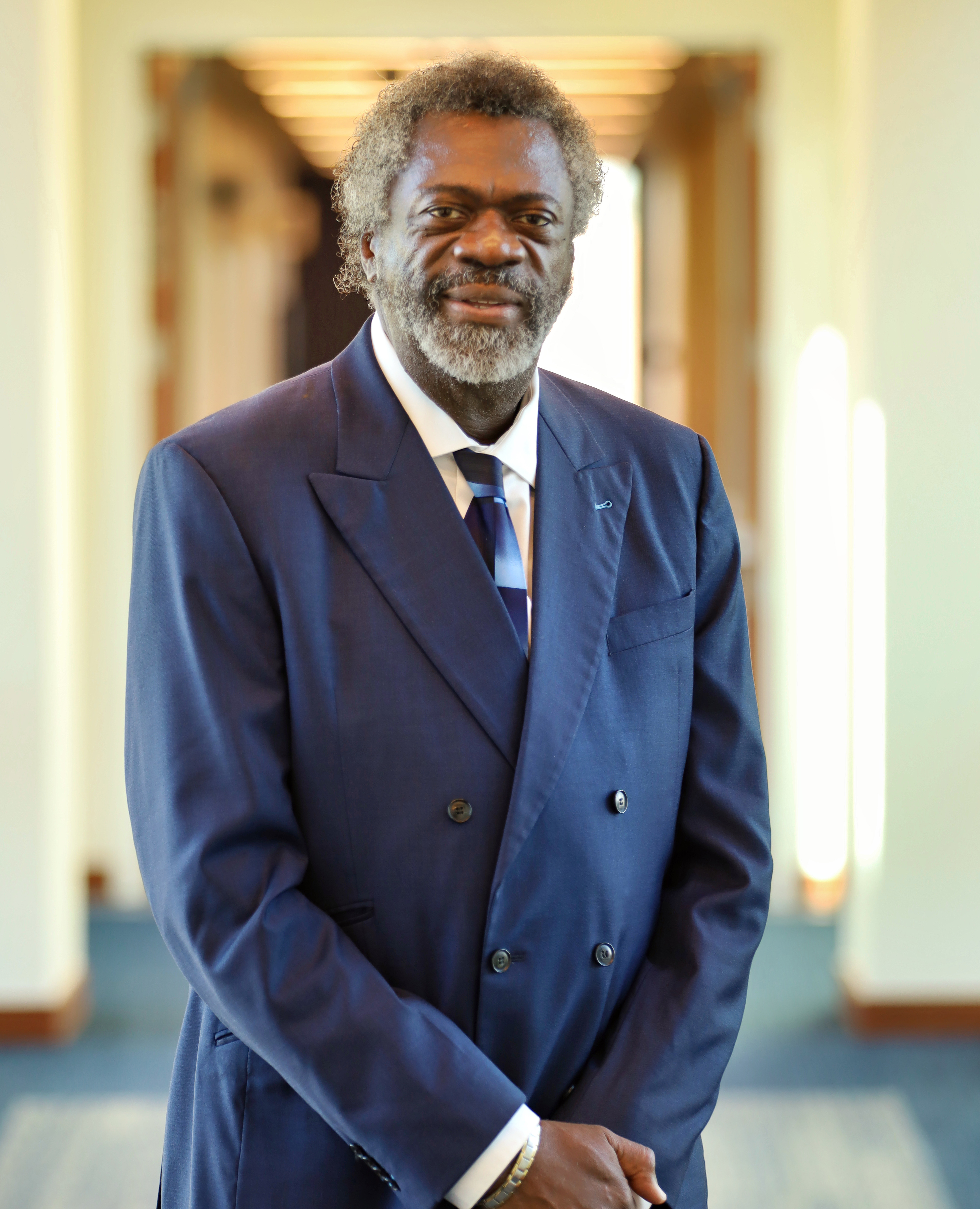 Ernest Grant stands in a well lit hallway, posing for a photo. He is wearing a navy blue suit.