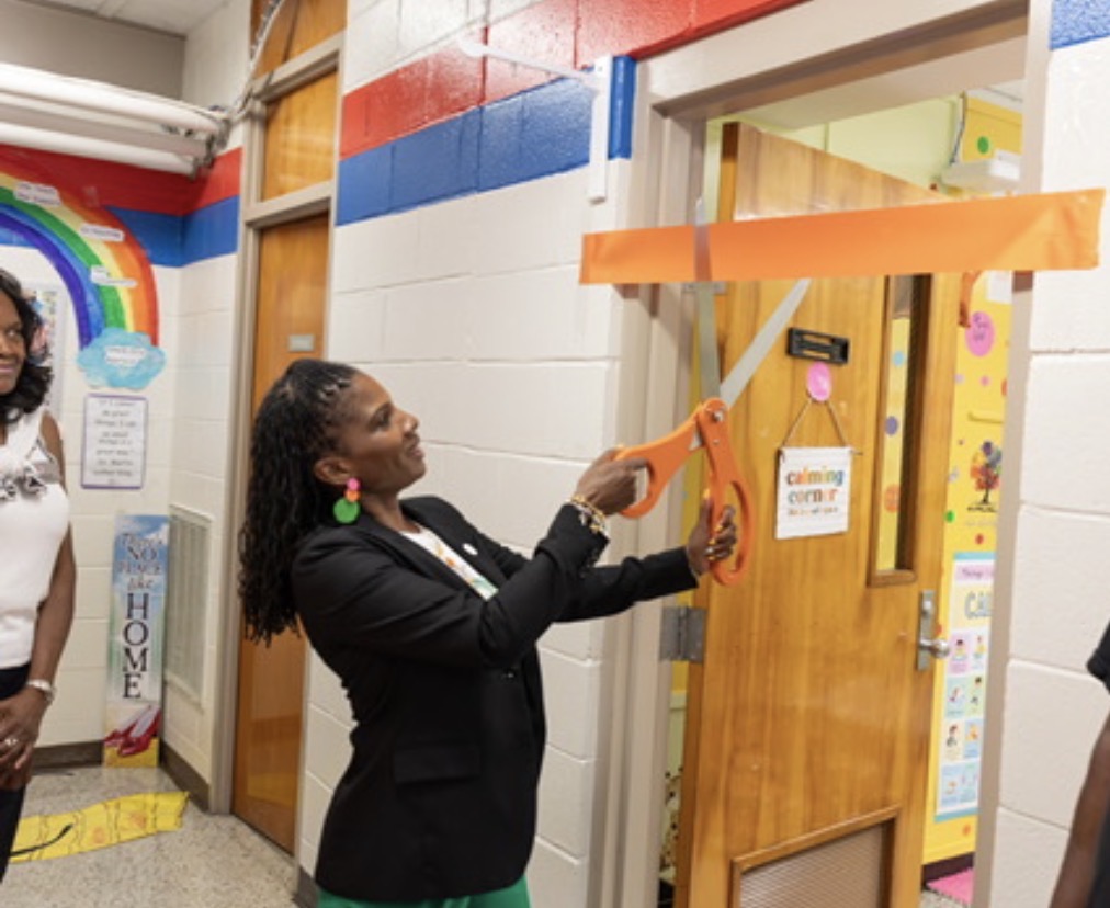 Lydia Nichols using giant scissors to cut the ribbon in front of a classroom door