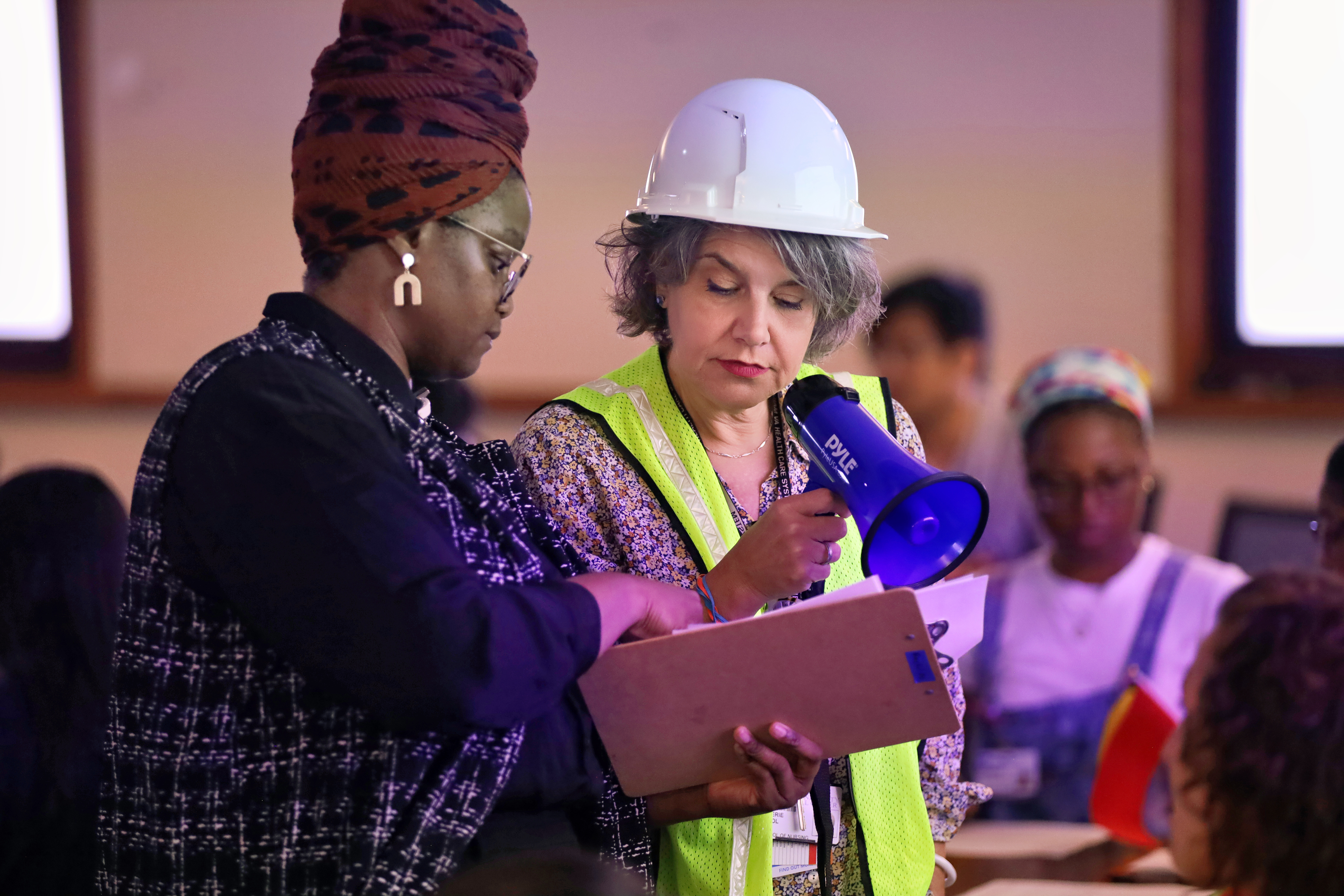 Two faculty lead a disaster simulation. One is wearing a white hard hat, yellow safety vest, and holds a bullhorn.