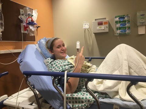 A person in a hospital bed smiles for the camera. They are in a hospital gown and have a blanket on their lap and are giving the peace sign.