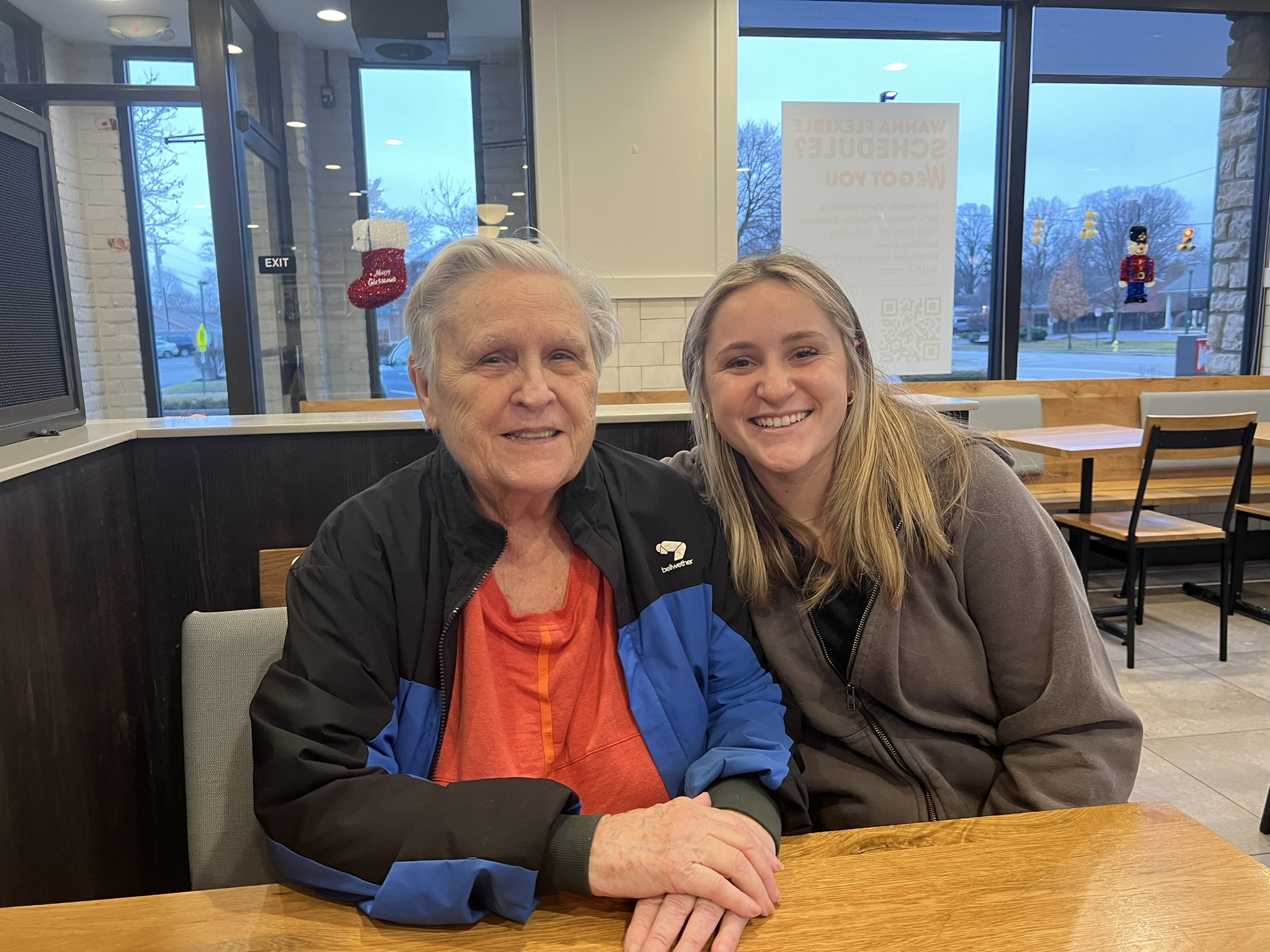 Two people pose for a photo. One is older and wearing a dark jacket over a light pink blouse, the other younger. They appear to be in a restaurant.