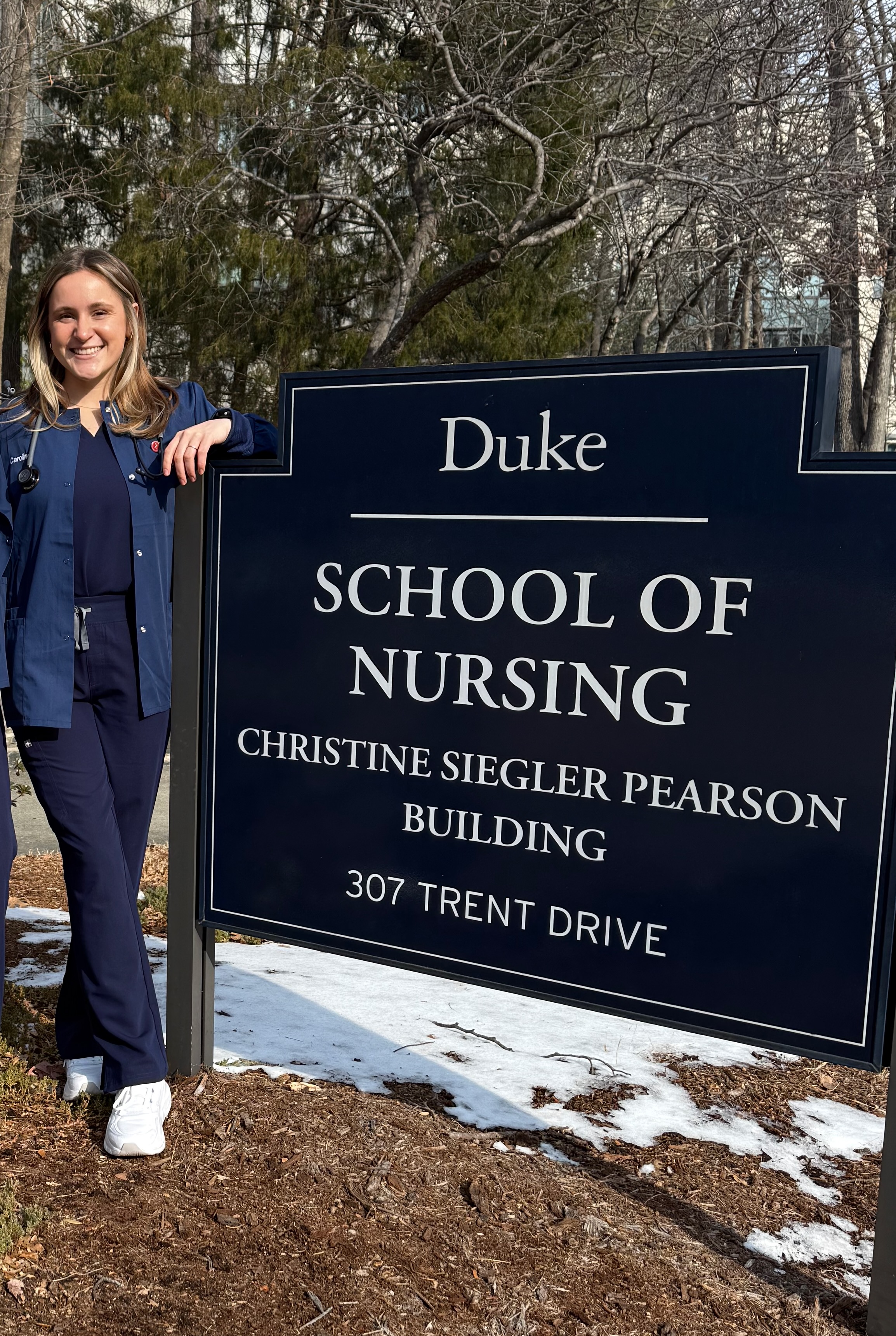 A nursing student poses in front of the sign for Duke University School of Nursing. They are wearing Duke Blue scrubs.