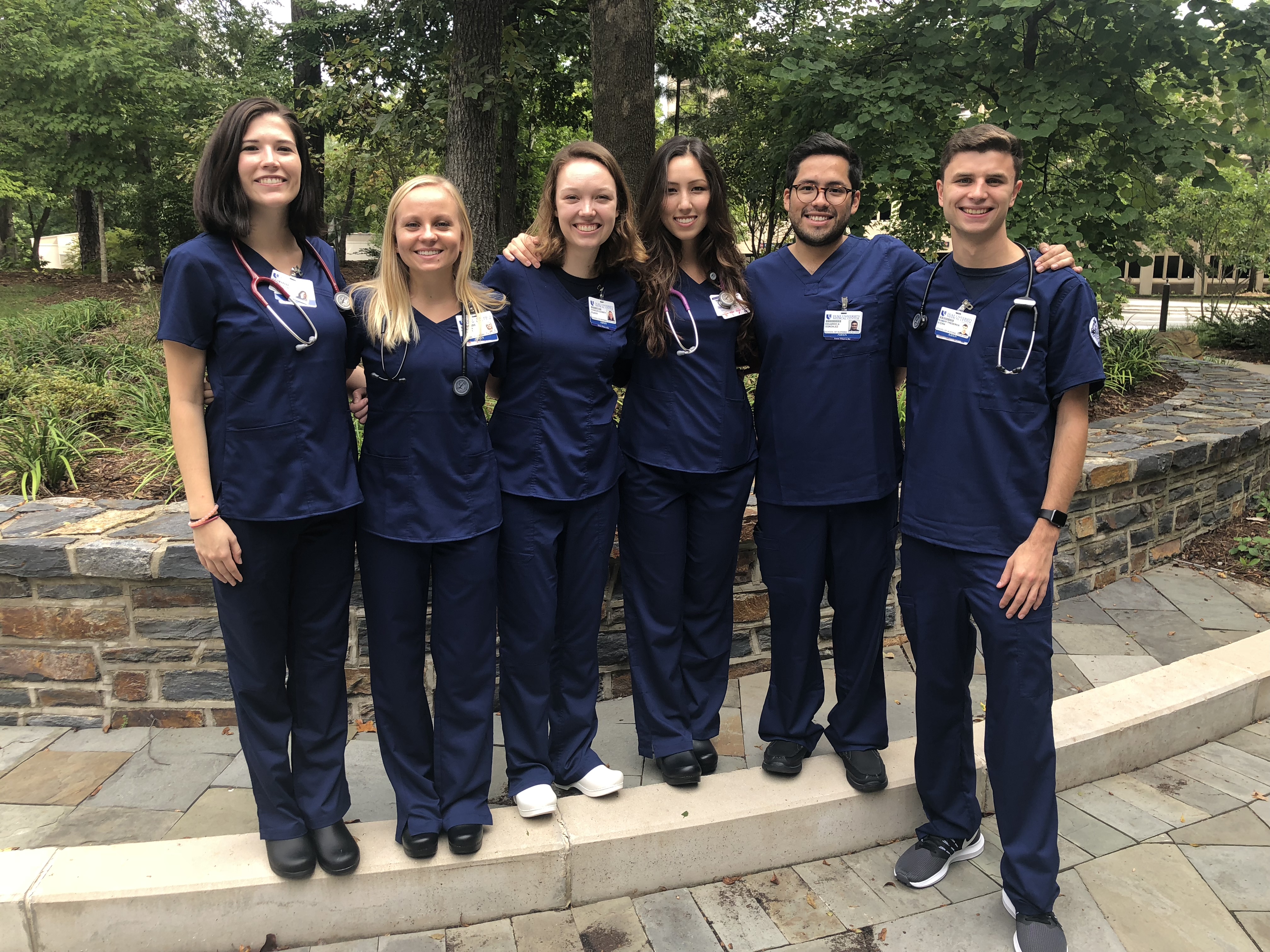Six people in blue scrubs standing in a courtyard with trees in the background