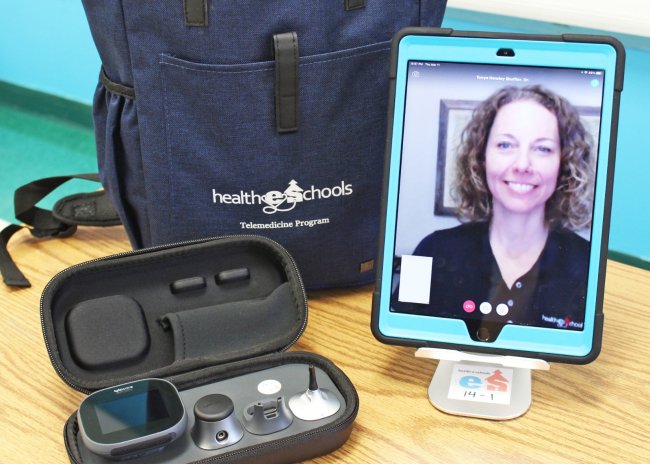 A nurse appears on a mobile screen as part of a telehealth visit. There is a backpack and audio equipment on the desk next to the tablet.