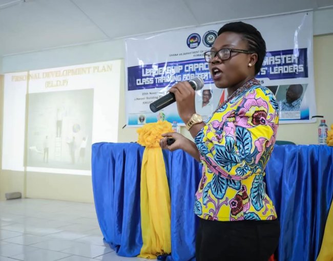 A woman in a brightly colored blouse stands at the front of a room, speaking into microphone that she is holding. 