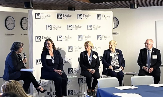 Terry McDonnell and Michael Relf sit at either end of a panel seated in front of a Duke in DC banner.