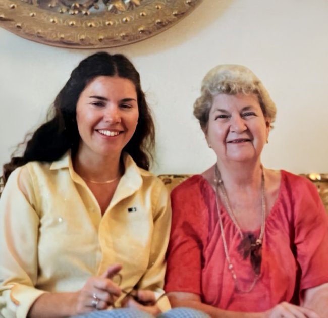 Two women pose for a photo. The younger woman on the left has long dark hair and is wearing a yellow blouse. The older woman on the right has short blonde hair and is wearing a pink blouse.