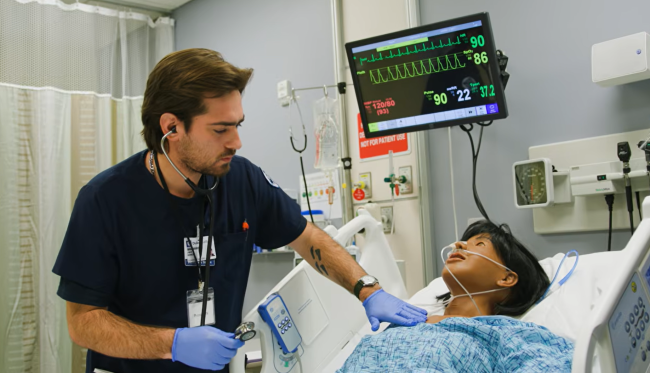 Jake Benoit in scrubs with gloves on examining a manekin in the Center for Nursing Discovery simulation lab