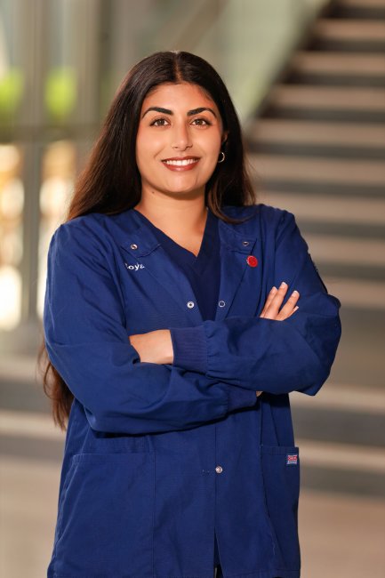 A person poses in a well-lit atrium. Their arms are crossed and they are wearing navy blue scrubs.