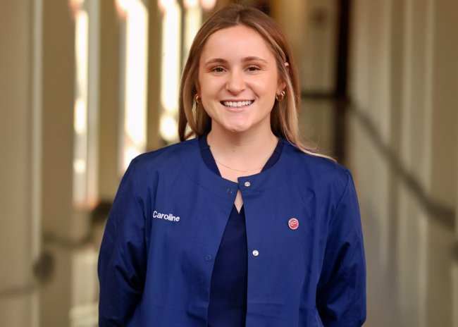 Caroline Bunker poses in a well-lit atrium. They are wearing Duke Blue scrubs.