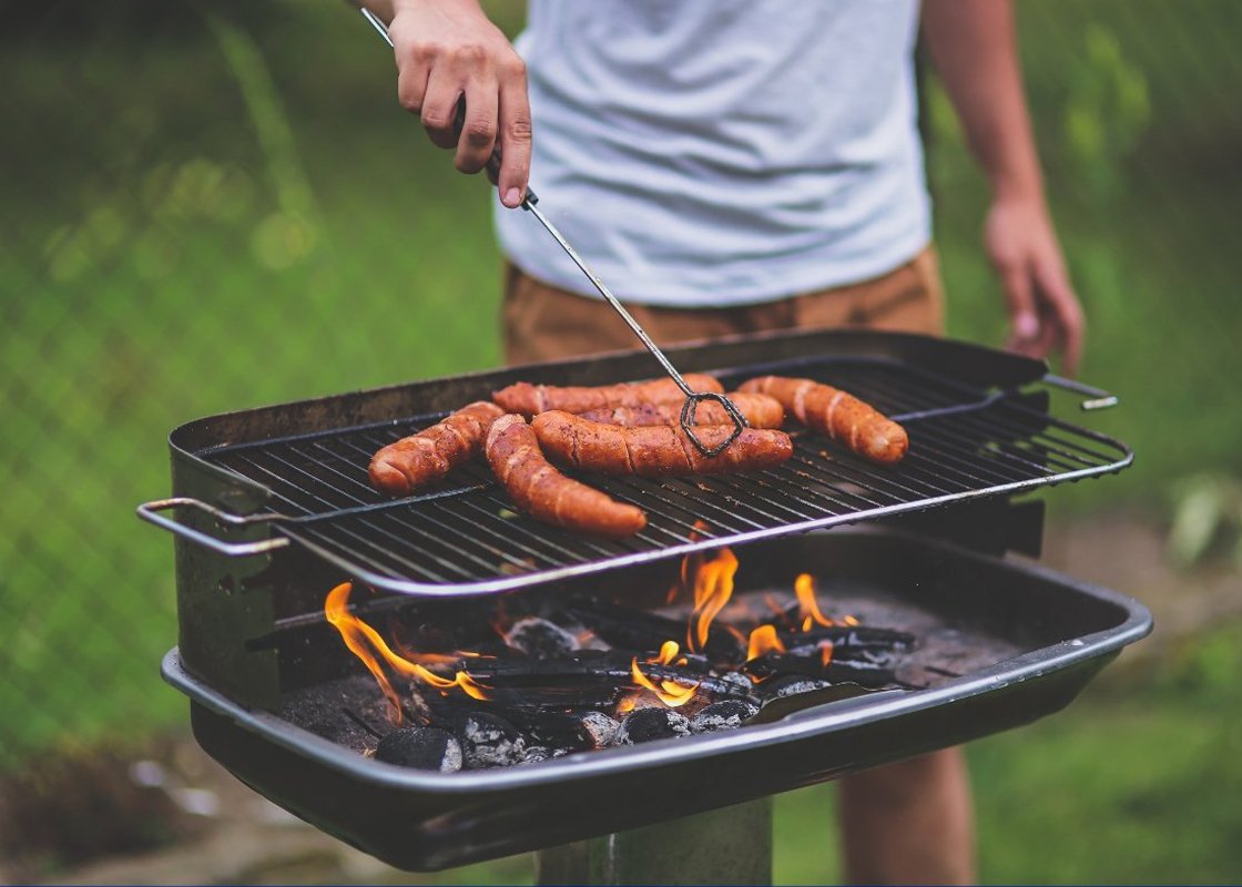 A person stands over a lit grill outside that has hot dogs or sausages on it.