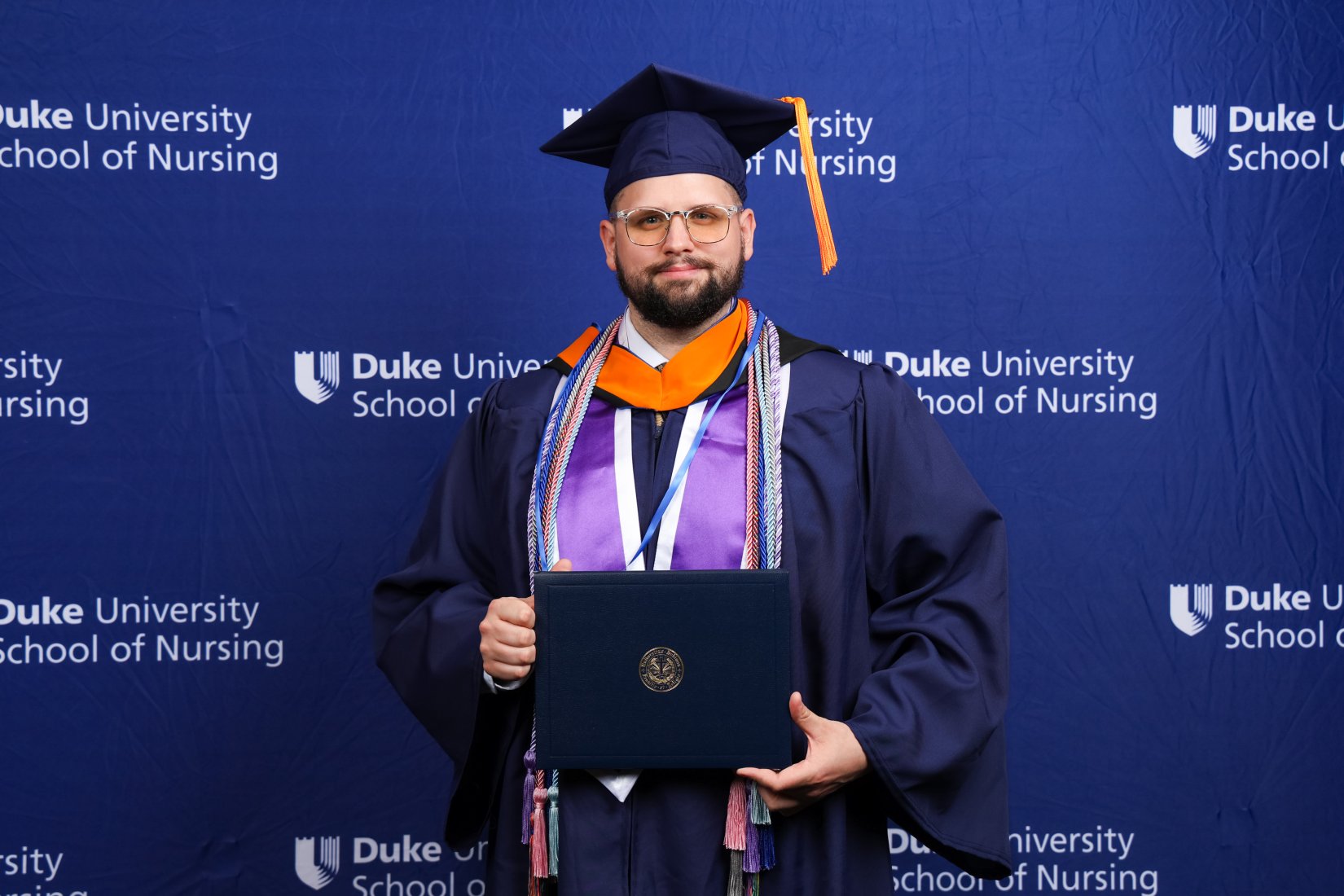 A student poses for a photo during graduation from Duke School of Nursing. He is wearing full regalia including many cords and is holding his diploma cover.