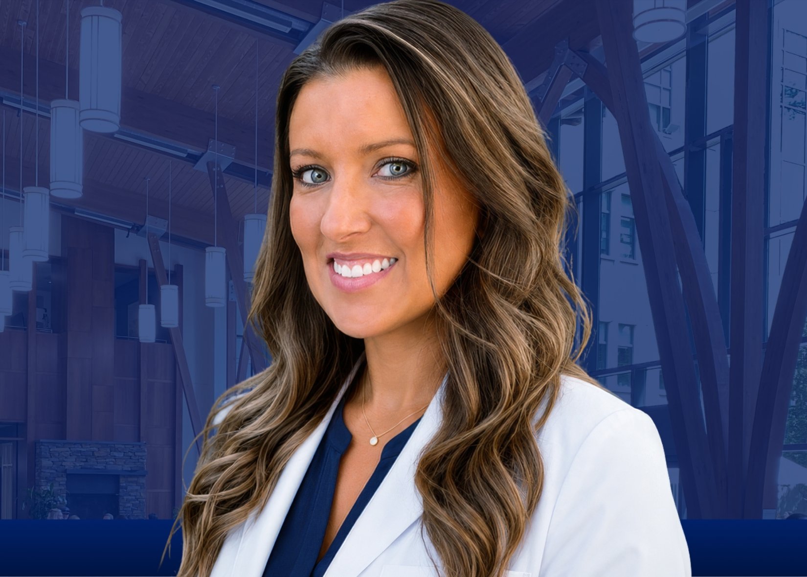 Ashley Batts poses for a headshot against a grey backdrop. They are wearing a white lab coat over navy blue scrubs, and have wavy long brown hair.